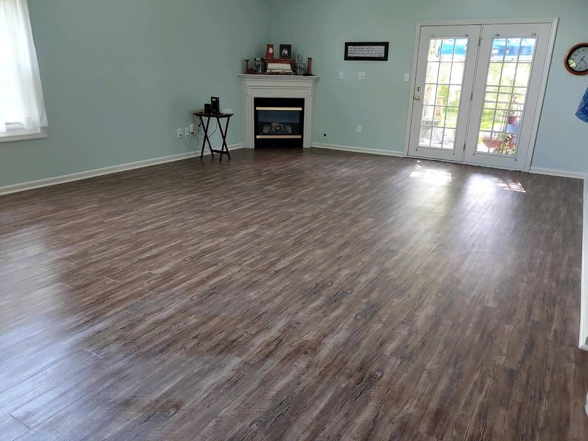 A large empty living room with a newly laid wide-plank floor running the length of the room, sage-green walls, a white fireplace and glazed double doors letting in daylight.
