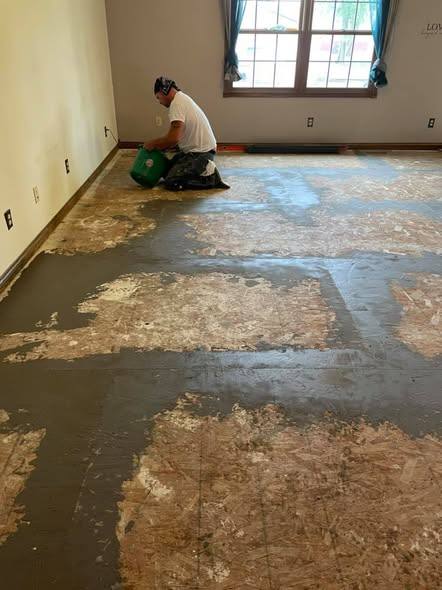 The same room a step further in, the carpet gone and the plywood subfloor patched and skim-coated flat, a worker kneeling by the window with a bucket.