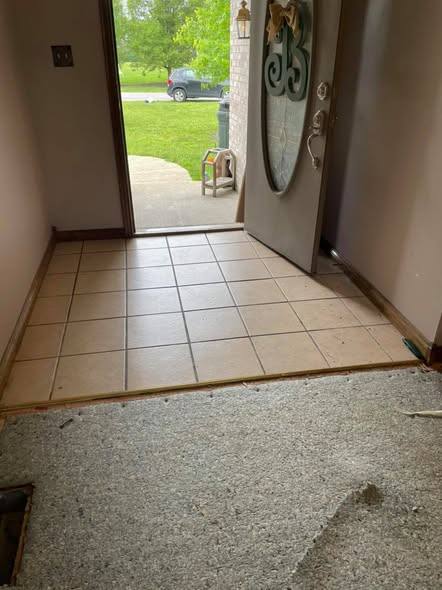 A house entry with the front door standing open onto a driveway, old beige floor tile in the doorway and the carpet beyond cut back to the underlay.