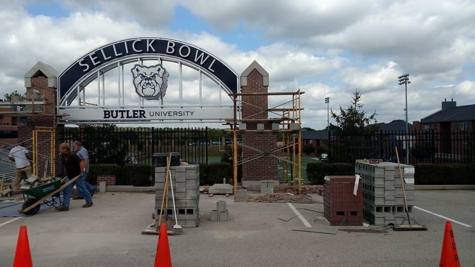 Two workers laying brick piers beside a wheelbarrow and stacked pallets of brick and block, beneath a steel arch sign reading Sellick Bowl, Butler University.